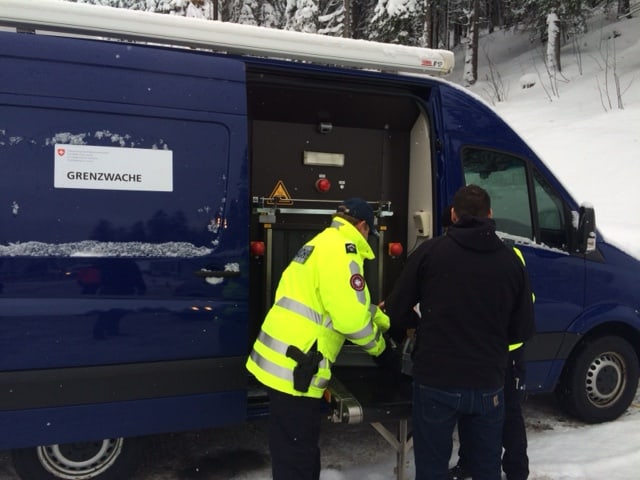 Ein Grenzwächter und ein Zivilist hieven ein Gepäckstück auf ein Förderband,