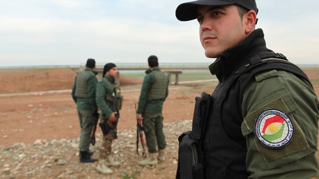 Vier irakisch-kurdische Kräfte, bewaffnet und in Uniform, stehen in der Nähe einer Brücke. 