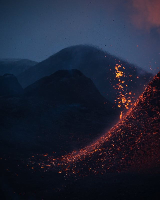 In Blautönen gefärbte Berge im Hintergrund, im Vordergrund sieht man, wie Lava aus einem Vulkan spritzt.