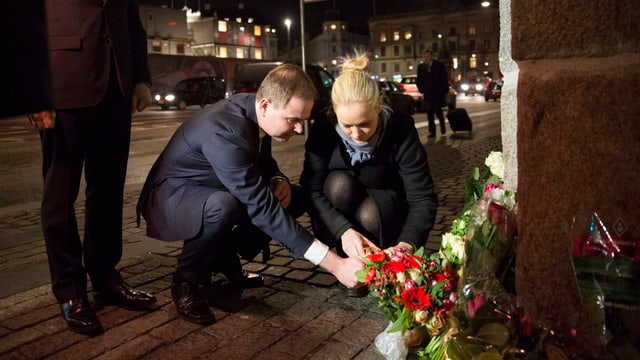 Eine Frau und ein Mann gehen in die Knie, um Kerzen anzuzünden. Daneben stehen ein paar Blumen.