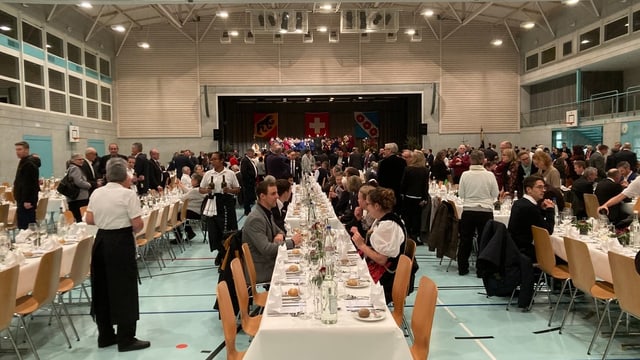 Drei Reihen Tische und Stühle in einer Mehrzweckhalle, Menschen strömen in den Saal.