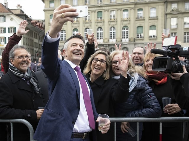 Ein Mann macht ein Selfie mit einem Handy, dahinter jubeln viele Menschen.