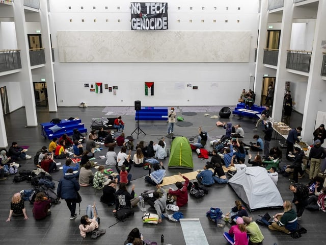 Demonstration in einer grossen Halle mit Menschen, die um Banner mit der Aufschrift 'NO TECH GENOCIDE' sitzen.