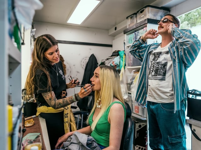 Eine Frau schminkt eine andere Frau, während ein Mann mit Sonnenbrille zuschaut.