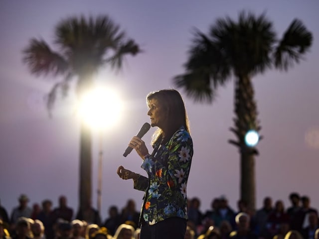 Nikki Haley steht auf der Bühne und spricht in Mikrophon. Im Hintergrund sind zwei Palmen. 