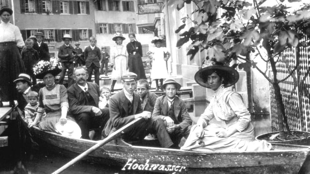 Gruppe von Menschen in einem Ruderboot mit dem Namen 'Hochwasser', Stadt im Hintergrund.