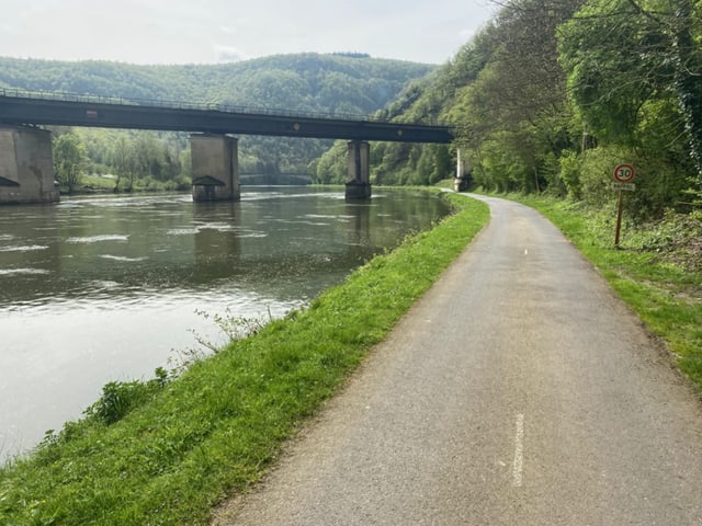 Radweg am Flussufer mit Brücke im Hintergrund.