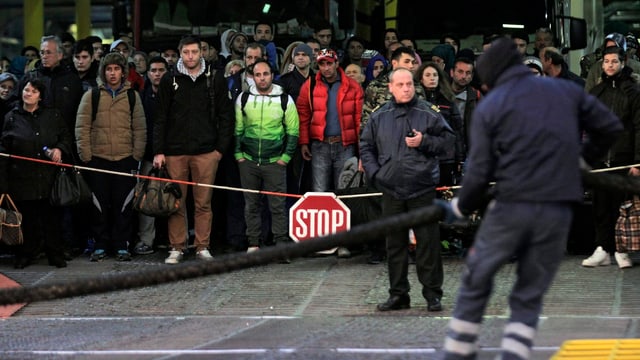 Zahlreiche Flüchtlinge stehen hinter einer Kette mit einem Stopp-Schild daran. Vorne zwei Beamte.