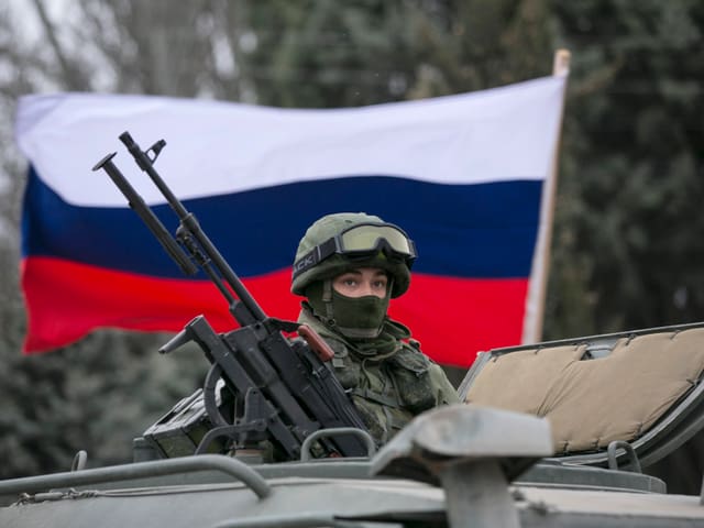 Ein schwer bewaffneter russischer Soldat auf einem Panzerfahrzeug, dahinter weht die russische Flagge. 