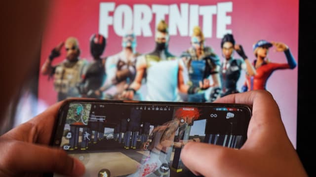 Jemand, der Fortnite auf einem Smartphone spielt, im Hintergrund Spiellogo und Charaktere. 