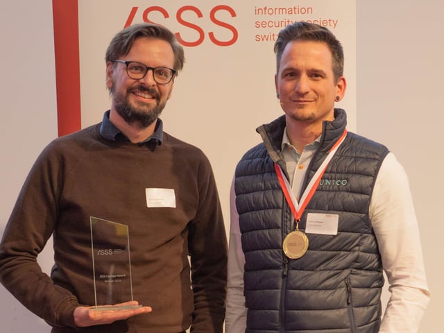 Zwei Männer, einer im braunen Pulli mit einem durchsichtigen Pokal, der andere mit einem blauen Gilet und einer Medallie