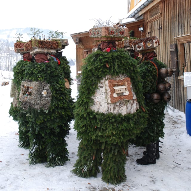 Mehrere mit Tannenzweigen und Hauben geschmückte Personen stehen im Schnee im Kreis.