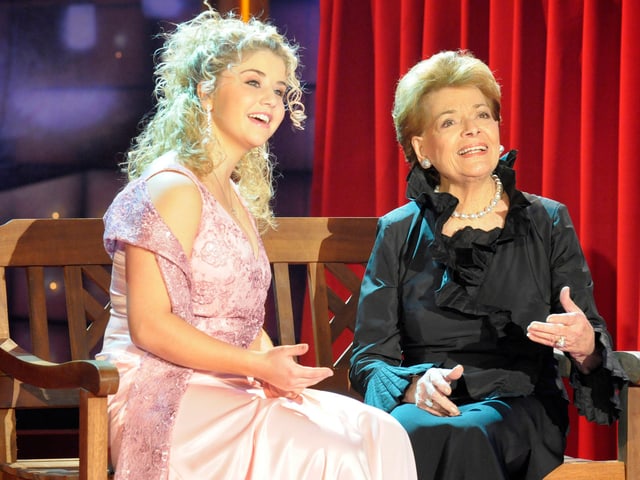 Beatrice Egli und Lys Assia sitzen vor einem roten Vorhang auf einer Holzbank.