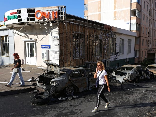 Verbrannte Autos vor zerstörtem Gebäude, Menschen gehen vorbei.