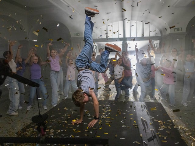 Eine Junge tanzt auf einem Tisch. Rundherum feuern ihn andere Tänzer:innen an. Goldenes Konfetti fliegt durch die Luft. 