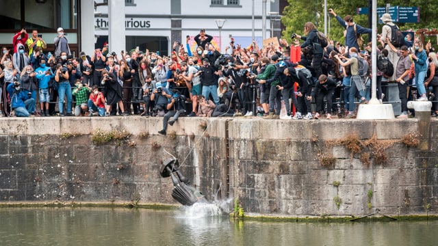 Statue wird ins Meer geworfen, Demonstranten sehen zu.