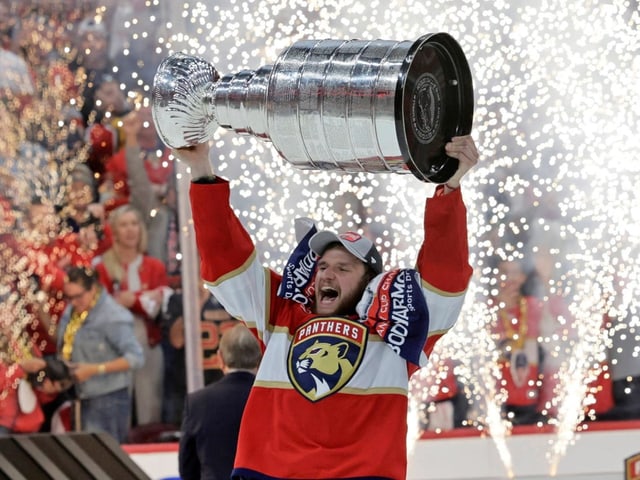 Eishockeyspieler hält Stanley Cup Trophäe hoch mit Funken im Hintergrund.