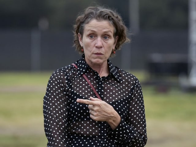 Mc Dormand looks at the camera and points to the left with his index finger.