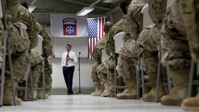 Soldaten sitzen in einer Halle auf Stühlen, vor ihnen spricht ein zivil gekleideter Mann.