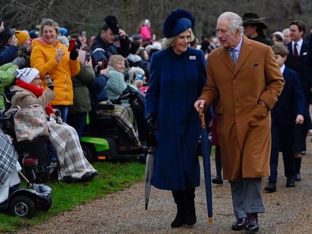 König Charles geht mit Camilla einen von Zuschauern flankierten Weg entlang.