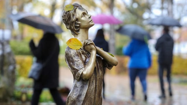 Eine Statue auf einem Friedhof, dahinter Menschen, die ein Grab besuchen.