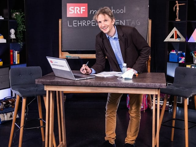 Raphael Labhart steht an einem Tisch mit zwei Stühlen in einem Schulzimer. Dahinter sind eine Wandtafel und Regale.