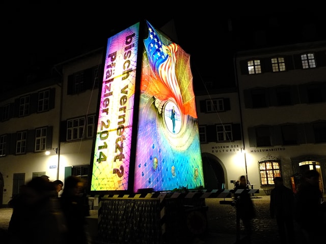 Alleinstehende Laterne der Pfälzler mit den Silhoutten von Betrachtern, dahinter erleuchtete Fenster.