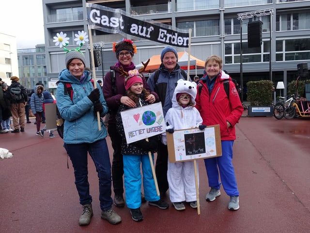 Drei-Generationen-Familie posiert mit Plakaten, zum Beispiel mit der Aufschrift «Gast auf Erden»