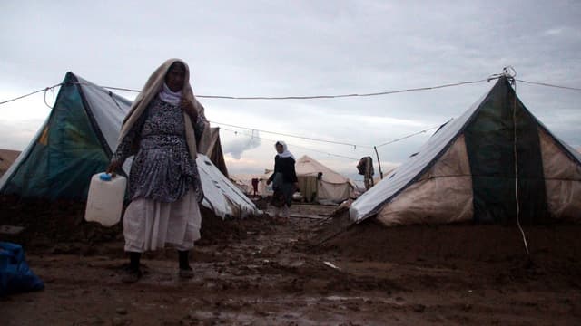 Eine Frau mit einem Wasserbehälter in der Hand läuft auf einem schlammigen Weg durch ein Flüchtlingslager