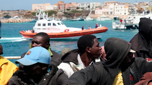 Mehrere Migranten aus Afrika im Vordergrund, im Hintergrund ein Boot der italienischen Küstenwache.