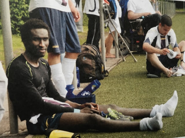 Ein schwarzer Mann sitzt neben dem Spielfeld auf dem Rasen und hat seine Fussballschuhe ausgezogen.