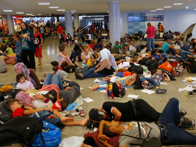 Zahlreiche Menschen sitzen und liegen auf dem Boden der Unterführung vor dem Bahnhof. Viele Kinder. Papiere liegen herum.