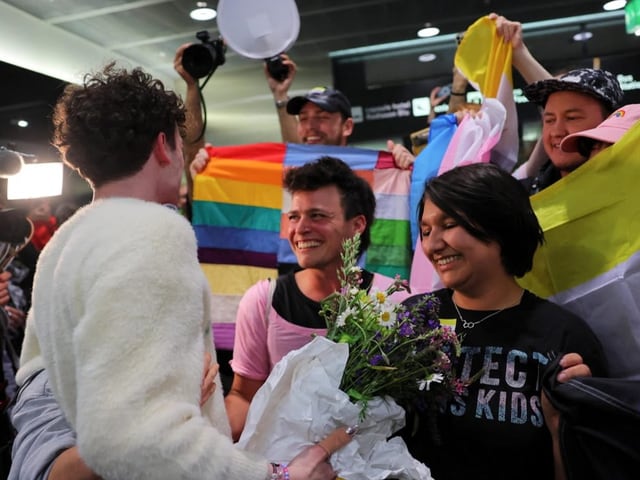 Freudige Begrüssung von Personen mit Regenbogenflaggen in einem belebten Raum.