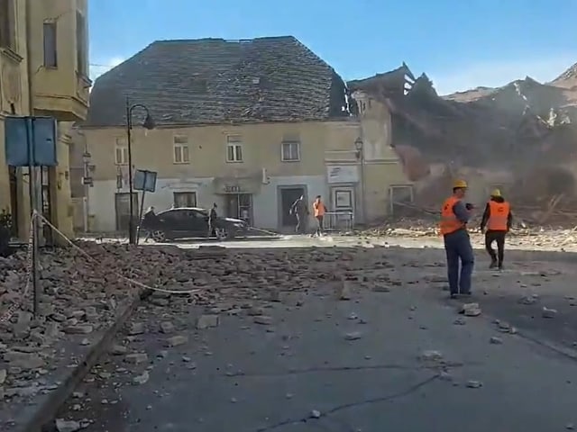 View of a wide street, rubble and debris on the ground, a heavily damaged house in the background.