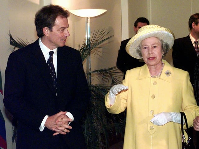 Tony Blair mit Anzug und Krawatte steht neben der Queen, die einen hellgelben Mantel und Hut trägt.