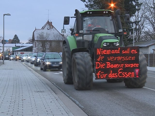 Ein Traktor fährt mit einem Plakat auf einer Strasse und verursacht Stau.