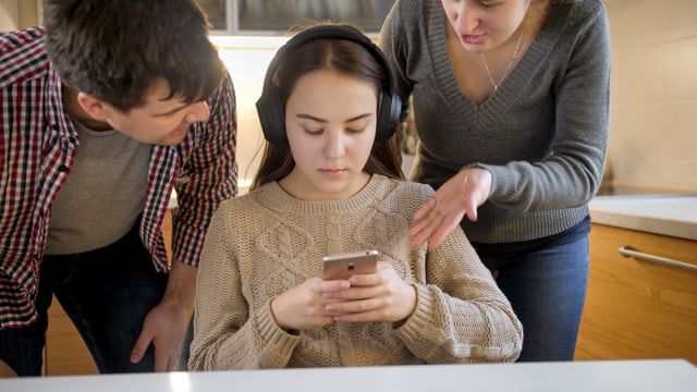 Ein Vater und eine Mutter reden auf ein in ihrer Mitte sitzendes Mädchen ein, das ein Smartphone in den Händen hält.