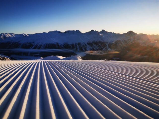 Frisch präparierte Skipiste im Vordergrund und verschneite im Berge im Hintergrund. Dazu blauer Himmel.