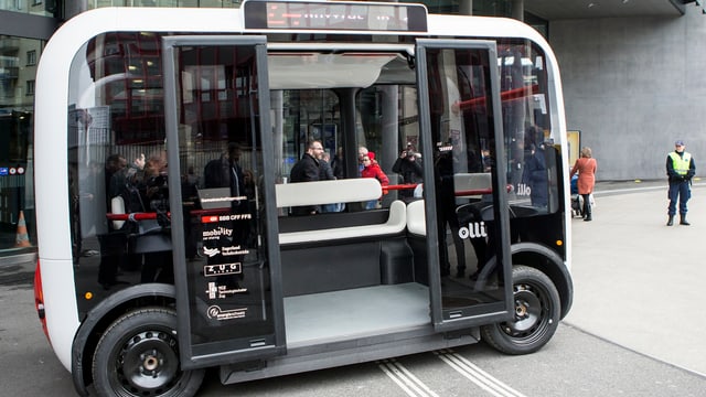 Rechteckiger Bus, der Platz bietet für ca. 10 Personen, steht vor dem Bahnhof Zug mit offen Türen. Daneben sehen zwei Menschen.