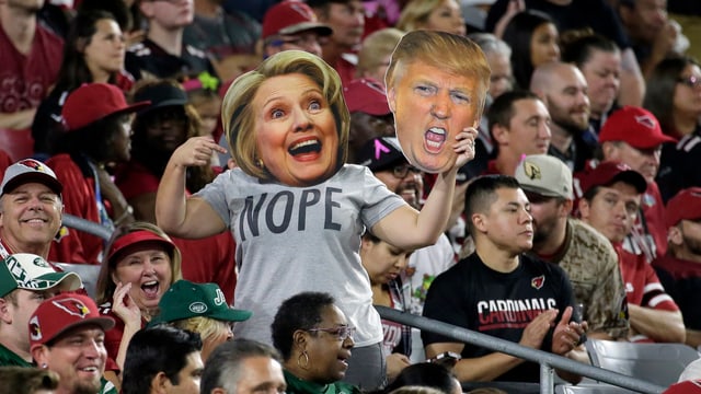 Jemand hält eine Clinton- und eine Trump-Maske in einem Stadion. Auf dem T-Shirt steht «Nope».