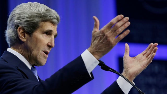 John Kerry an einem Mikrofon, er streckt beide Arme aus.