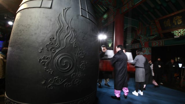 A metal bell the size of a man rings.  Four people move the wooden stick that rings the bell.