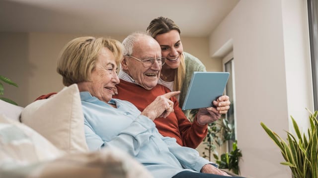 Älteres Paar und junge Frau betrachten gemeinsam ein Tablet auf dem Sofa.