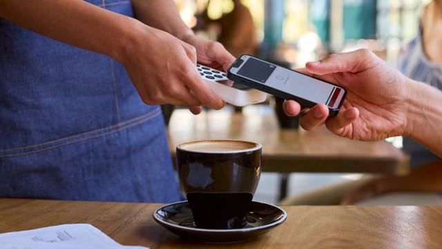 Hände, zwei halten Lesegerät, eine rechts hält Handy zum Bezahlen. Auf dem Tisch eine Tasse Kaffee.