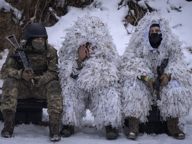 Drei Männer sitzen in Tarnanzügen und Waffen.