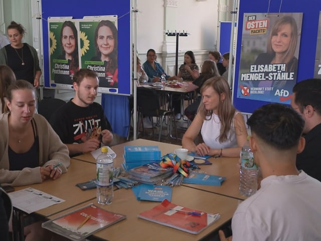 Menschen sitzen an einem Tisch mit Wahlkampfbroschüren, im Hintergrund politische Plakate.