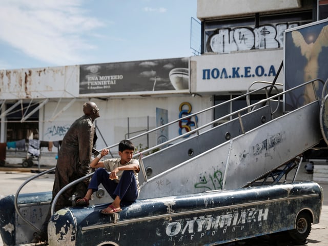 Ein Junge sitzt neben einer Statue auf einem alten Gangway auf Athens stillgelegtem Flughafen. 