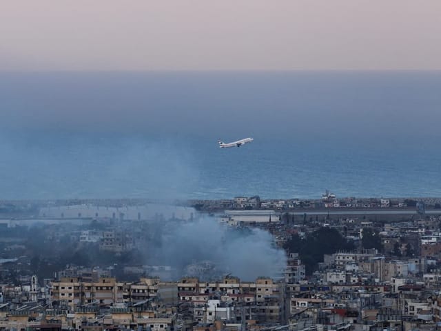 Flugzeug startet über Küstenstadt, Meer im Hintergrund.