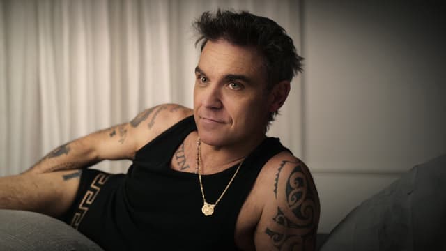 Robbie Williams in schwarzem Tanktop und schwarzen Boxershorts auf dem Bett