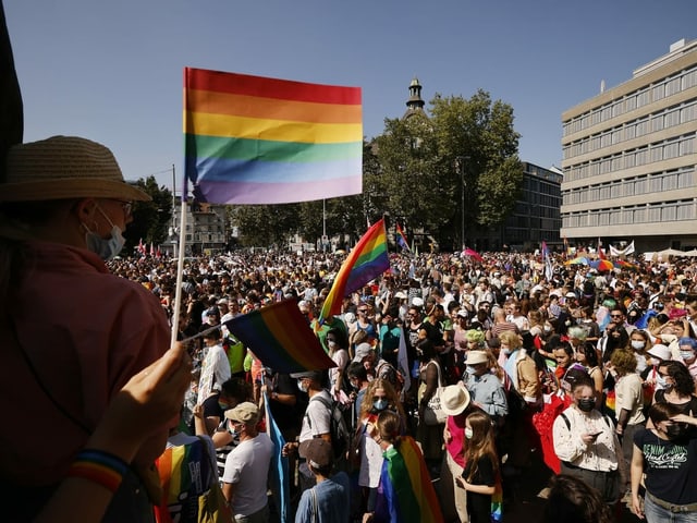 Menschen mit Regenbogenfahnen stehen auf einem Platz.